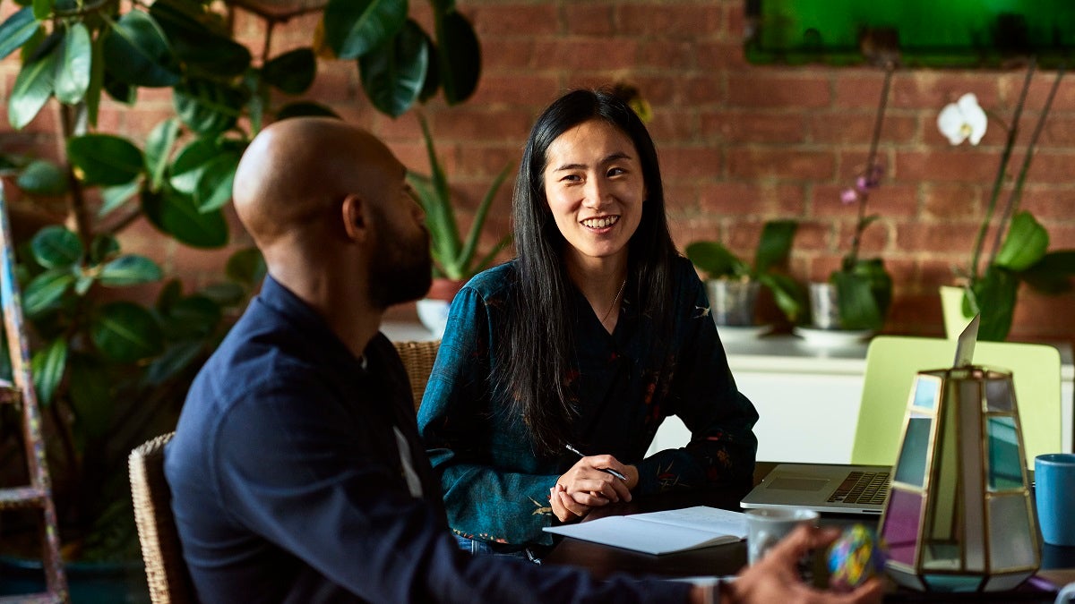 Two creative designers sitting in meeting in modern office, woman with note paper and pen listening to man with cheerful expression