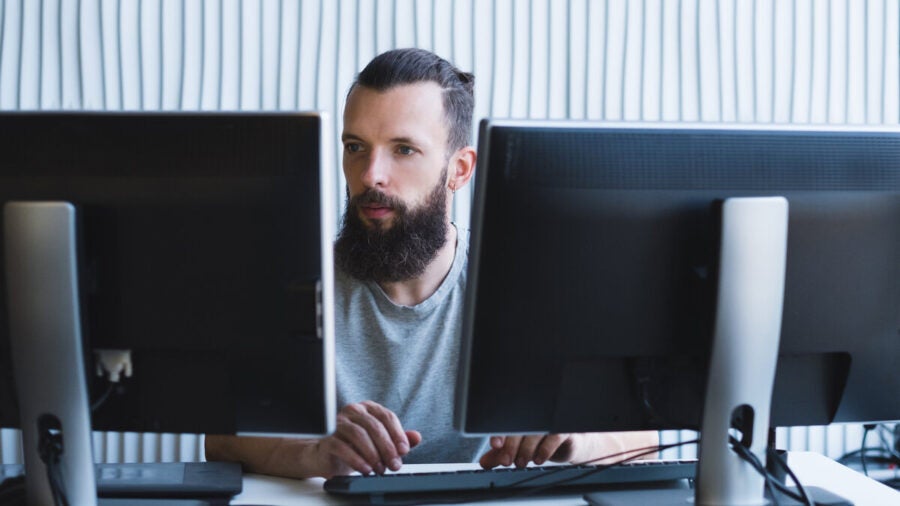 A man sits at an office desk between two computer monitors